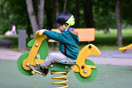 Boy bouncing on spring rocker shaped like a motorcycle in playground, wearing helmet and green hoodie. Joyful scene captures movement, excitement, and childhood playfulness in colorful outdoor space.の写真素材
