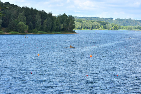 Two athletes row a boat on a calm lake during a training session or race. Forested shoreline and lane buoys frame the peaceful water scene.のeditorial素材
