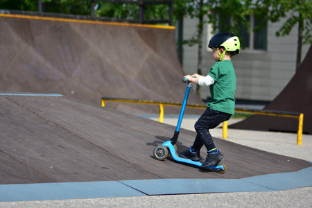 Sporty toddler boy wearing green shirt and helmet rides blue three-wheel scooter at skate park ramps...