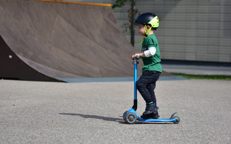 Young boy wearing a black helmet rides a blue three-wheeled scooter at a skate park, concentrating...
