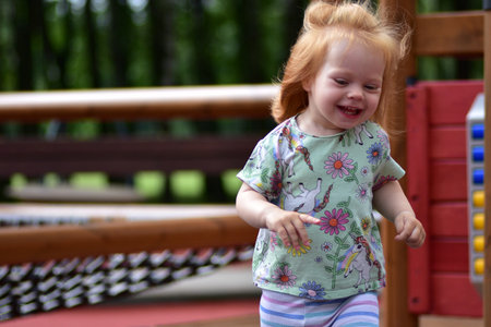 Smiling red-haired toddler in unicorn shirt and striped leggings runs on wooden playground in park. Joyful childhood and active outdoor play.の写真素材