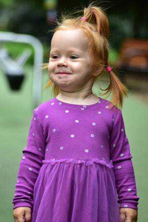 Close-up portrait of toddler girl with red hair tied in ponytails, wearing purple dress. Cute child outdoors in playground setting, expressing thoughtful look.の写真素材