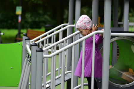 Little girl in purple dress and hat holding railing on playground bridge. Toddler carefully...