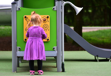 Little girl with red hair in ponytails wearing purple dress standing at yellow playground activity board. Toddler turning knobs and learning through outdoor play.の写真素材