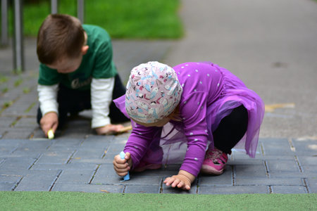 Two young children drawing with colorful chalk on pavement outdoors. A girl in purple dress and a boy in green shirt concentrate on creative play in the park. Childhood activity concept.の写真素材