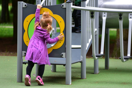 Cute toddler girl in purple dress climbing into playground entrance with yellow frame. Child...