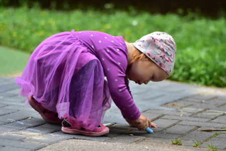Small girl in purple dress and hat bending down on pavement and drawing with blue chalk. Child engaged in creative outdoor play, enjoying childhood fun and imagination.の写真素材