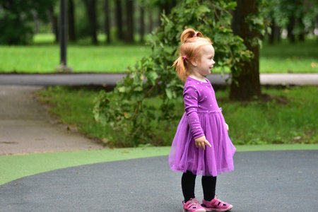 Little girl in purple dress smiling while standing on playground. Toddler enjoying summer playtime outdoors, expressing joy and happiness in childhood.の写真素材