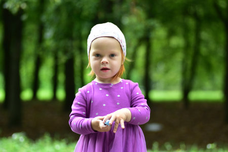 Cute toddler girl in purple dress and patterned hat holding blue chalk in her hands. Child stands in green park background, enjoying outdoor creative activity in summer.の写真素材