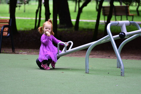 Little girl with red hair in purple dress sitting on playground seesaw, holding snack in hand. Toddler enjoys playful outdoor break during summer day.の写真素材