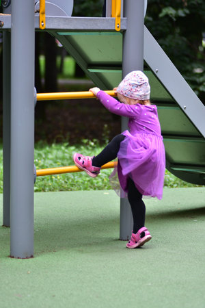 Little girl in purple dress climbing up yellow ladder at playground. Toddler enjoying outdoor...
