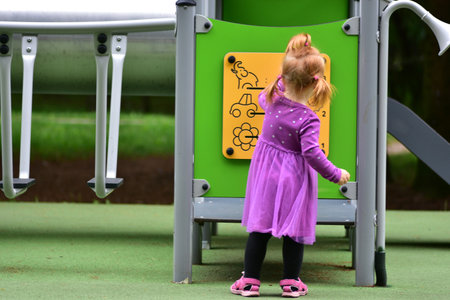 Little girl with red hair in ponytails wearing purple dress standing at yellow playground activity board. Toddler turning knobs and learning through outdoor play.の写真素材