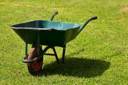 Old green metal wheelbarrow with red wheel standing on grass in sunlight. Traditional farm and gardening tool used for carrying soil and crops.の写真素材