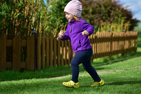 Little girl in purple jacket, pink hat and yellow shoes walks across garden path on sunny day. Cute child exploring outdoors in natural rural environment.の写真素材