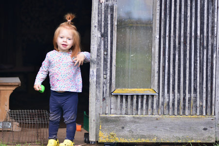 Cute red-haired toddler in floral shirt and yellow shoes standing by old wooden shed door, holding toy ball. Rustic countryside lifestyle moment with child outdoors.の写真素材
