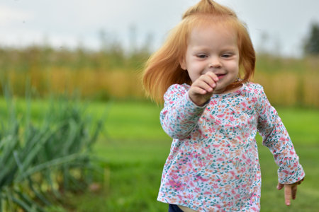 Red-haired toddler in floral shirt running happily on grass in countryside. Active child enjoying outdoor fun and freedom in rural summer nature.の写真素材