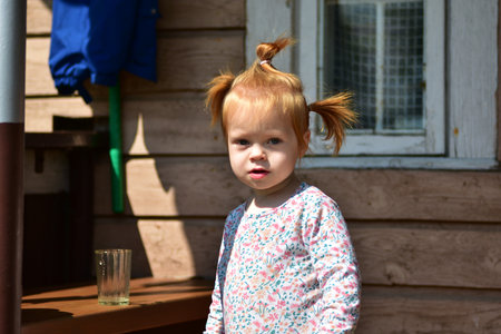 Little red-haired girl with two ponytails stands outside in front of wooden house wall. Childhood portrait with playful hairstyle in countryside environment.の写真素材