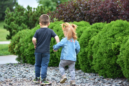 Adorable toddler girl with red hair holding hands with little boy as they walk together on gravel path by green bushes. Cute siblings enjoying friendship, bonding, and carefree childhood outdoors.の写真素材