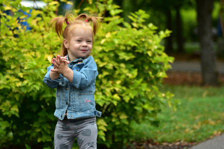Cute toddler girl with pigtails wearing denim jacket standing near plants in playground. Child outdoors enjoying childhood curiosity and play.の写真素材