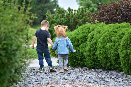 Adorable toddler girl with red hair holding hands with little boy as they walk together on gravel path by green bushes. Cute siblings enjoying friendship, bonding, and carefree childhood outdoors.の写真素材