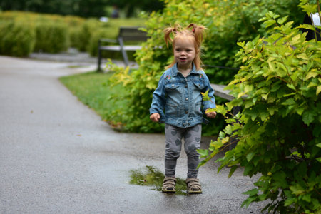 Cute toddler girl in denim jacket and heart leggings standing in a puddle on park path. Child enjoying playful happy moments splashing water after rain, exploring nature with joy.の写真素材