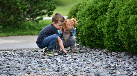 Little boy and red-haired toddler girl crouching together, exploring gravel stones in park. Children learning, curiosity, friendship, happy outdoor play, and childhood discovery moments.の写真素材