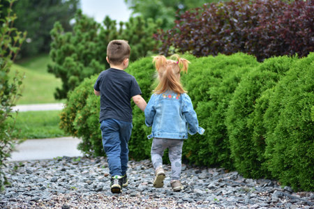 Adorable toddler girl with red hair holding hands with little boy as they walk together on gravel path by green bushes. Cute siblings enjoying friendship, bonding, and carefree childhood outdoors.の写真素材