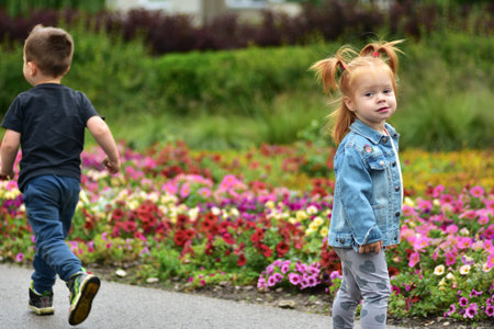 Cute toddler girl with red hair standing near colorful flower garden while boy walks away. Children outdoors in park, enjoying nature, carefree fun, and happy childhood moments.の写真素材