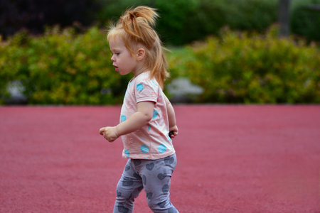 Cute red-haired toddler girl with pigtails running across red playground ground. Child enjoying fun movement, carefree outdoor play, and joyful moments of childhood.の写真素材