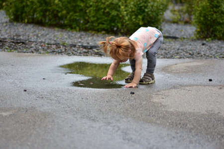 Little red-haired toddler girl bending over wet pavement to touch puddle water. Child exploring nature outdoors, enjoying fun splashing, curiosity, and happy playtime moments after rain.の写真素材
