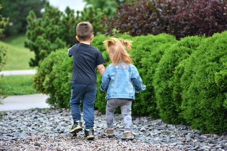Adorable toddler girl with red hair holding hands with little boy as they walk together on gravel path by green bushes. Cute siblings enjoying friendship, bonding, and carefree childhood outdoors.の写真素材