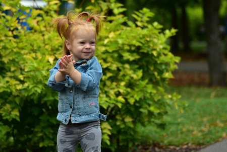 Cute toddler girl with pigtails wearing denim jacket standing near plants in playground. Child outdoors enjoying childhood curiosity and play.の写真素材