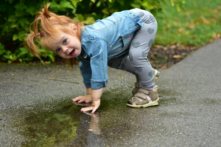 Red-haired toddler girl in denim jacket and heart leggings crouching to touch a puddle on wet pavement. Child exploring outdoors, enjoying happy moments splashing water after rain.の写真素材