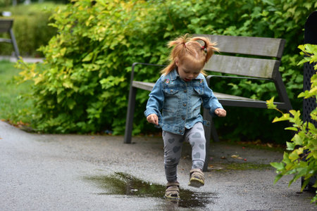 Cute toddler girl in denim jacket and heart leggings standing in a puddle on park path. Child enjoying playful happy moments splashing water after rain, exploring nature with joy.の写真素材