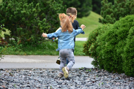 Red-haired toddler girl with pigtails chasing boy in black shirt on gravel park path. Children running, laughing, and enjoying carefree outdoor fun and happy childhood moments together.の写真素材