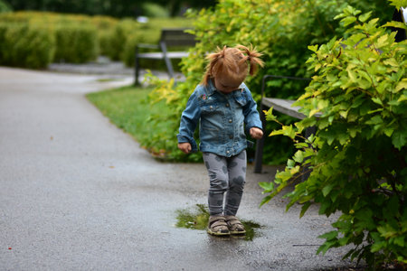 Cute toddler girl in denim jacket and heart leggings standing in a puddle on park path. Child enjoying playful happy moments splashing water after rain, exploring nature with joy.の写真素材