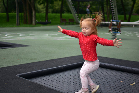 Little girl on trampoline jumping with arms stretched wide, wearing red shirt, playful energy and joyful outdoor movement captured in park playground.の写真素材