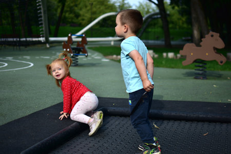 Toddler girl sitting on trampoline edge looking back smiling while boy stands beside holding feather. Fun childhood moment outdoors at modern playground in green park.の写真素材