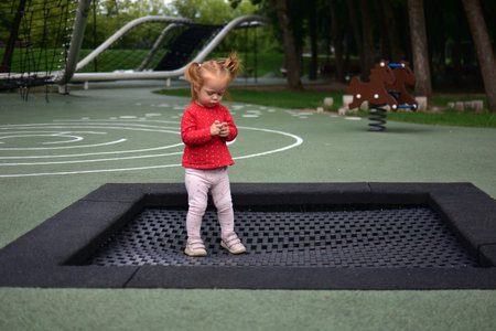 Little toddler girl standing on trampoline examining something in hands, focused moment outdoors at park playground with creative markings and green surroundings.の写真素材