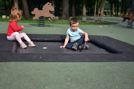 Boy sitting on trampoline area playing with feather while girl sits beside on edge, outdoor calm activity moment in park children zone with modern playground design.の写真素材