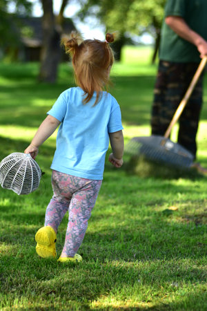 Red-haired toddler walking on green grass holding basket while grandfather rakes nearby. Outdoor play, countryside lifestyle, summer day family activities in nature.の写真素材