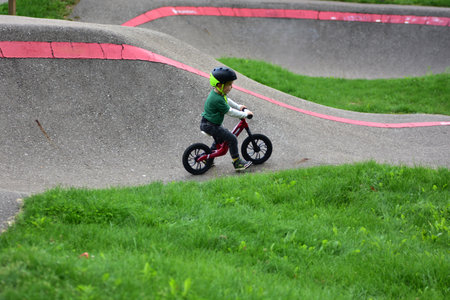 Toddler wearing a helmet rides a balance bike on an outdoor pump track. Child enjoying active play and developing balance and coordination skills in a green park environment.の写真素材