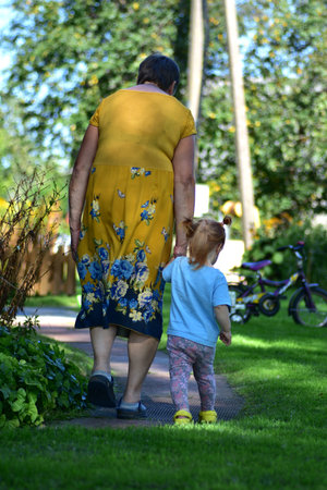 Elderly woman in a yellow dress with blue floral pattern walking hand in hand with a toddler girl in a blue shirt and patterned leggings, peaceful countryside family scene.の写真素材
