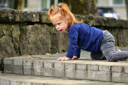 Determined red-haired toddler girl climbing outdoor stone stairs. Active child wearing blue shirt and grey jeans exploring independence and physical development.の写真素材