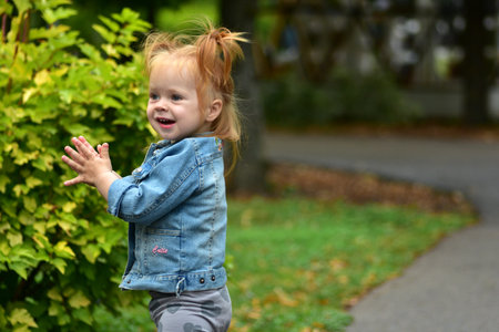 Cute toddler girl with pigtails wearing denim jacket standing near plants in playground. Child outdoors enjoying childhood curiosity and play.の写真素材