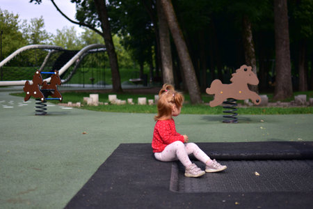 Toddler girl sitting alone on trampoline edge looking away, peaceful and calm moment at outdoor modern playground surrounded by trees and playful equipment.の写真素材
