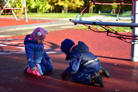 Two children in colorful overalls laughing and playing together on playground surface on cold autumn morning. Joyful childhood friendship moment. Siblings having fun outside, exploring natureの写真素材