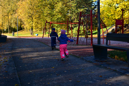 Two children in warm overalls running toward playground with swings and climbing frames on sunny autumn day, active childhood moment. Brother and sister playing together in empty autumn parkの写真素材