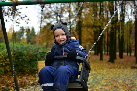 Cute toddler enjoying gentle swinging in park during autumn, smiling and dressed warmly for chilly...