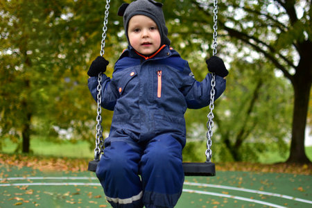 Preschool boy in blue snowsuit swinging outdoors on metal chains in park surrounded by autumn trees...